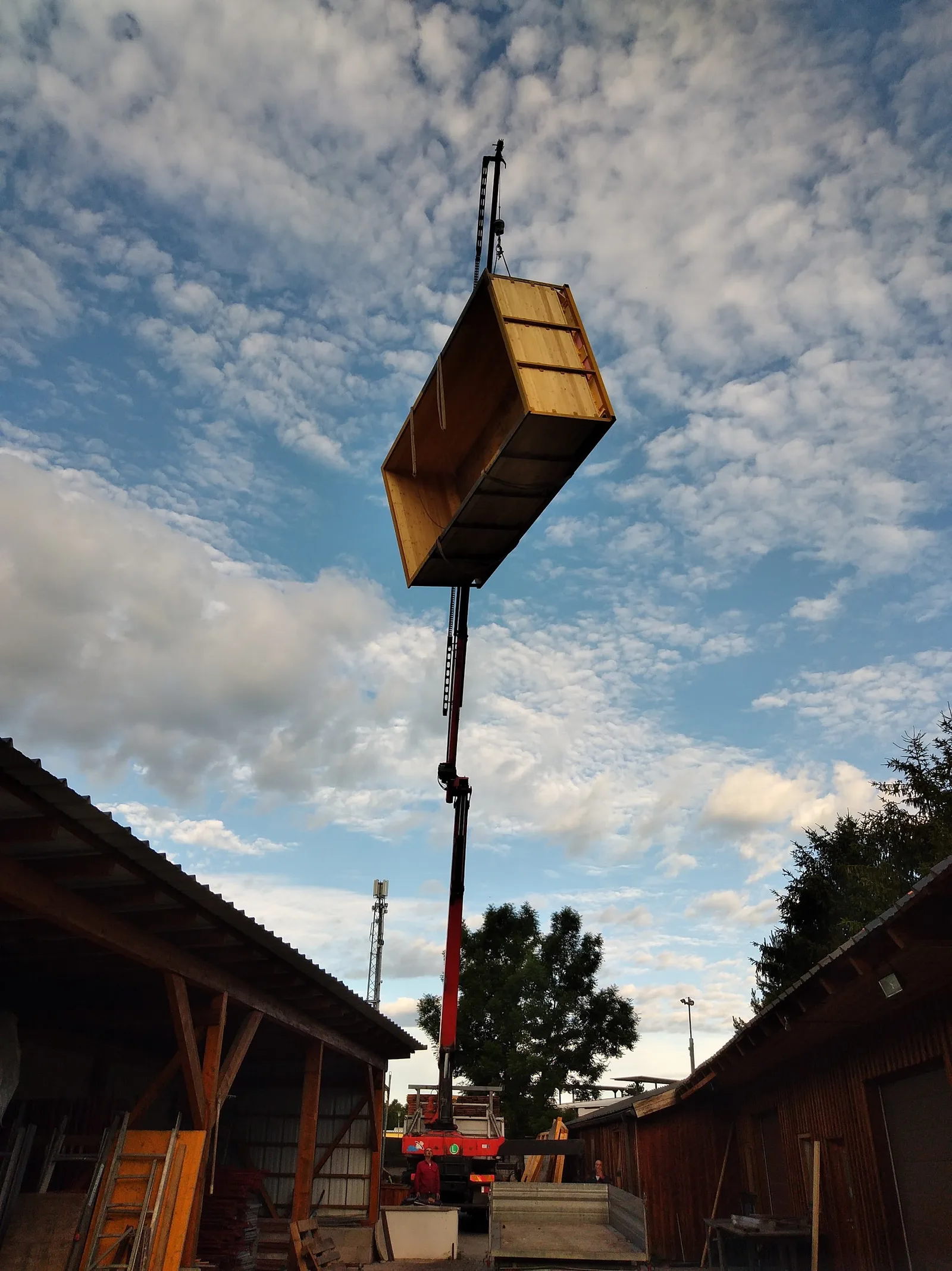 Ein Ladekran hebt eine Last hoch über einen Hof mit offenen Schuppen